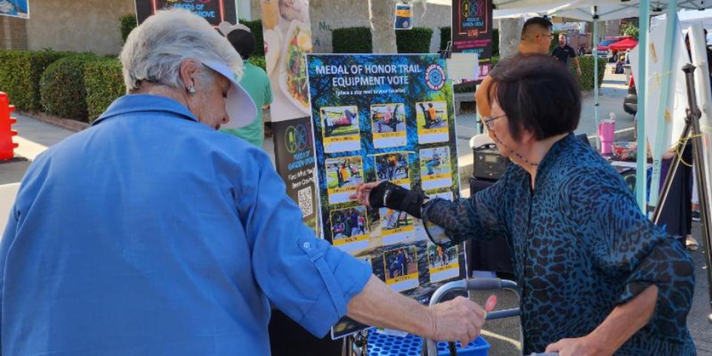 Two older women stand at an outdoor community event booth, reviewing a poster board titled "Medal of Honor Trail Equipment Vote." The board displays a grid of photos showing various outdoor fitness equipment options, each with a star-rating system for residents to vote on their preferred choices. A "Foods of Garden Grove" banner and a "Good Food GG Live" sign are visible in the background.