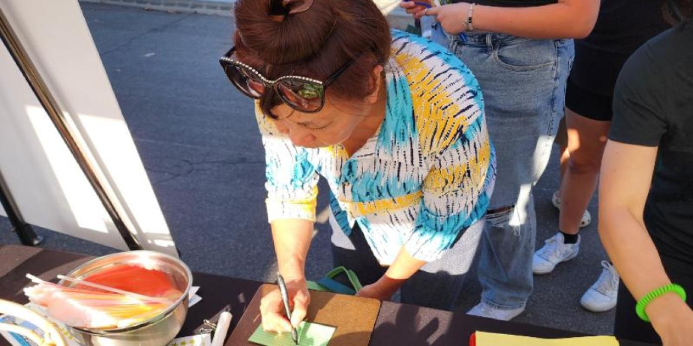 An overhead view of a woman leaning over a table at an outdoor community event, writing on a small green card. Art supplies, including a bowl of red material and scattered papers in various colors, are spread across the table. Other attendees are visible in the background.