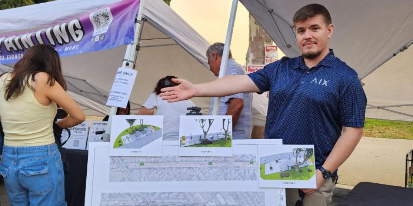 A young man in a navy blue polo shirt stands at an outdoor community event booth, presenting large printed site plan renderings for the Medal of Honor Trail fitness station installations. Three individual station layout plans are displayed in front of him, along with a wide overall trail map. A tent with a "Painting" banner is visible in the background.