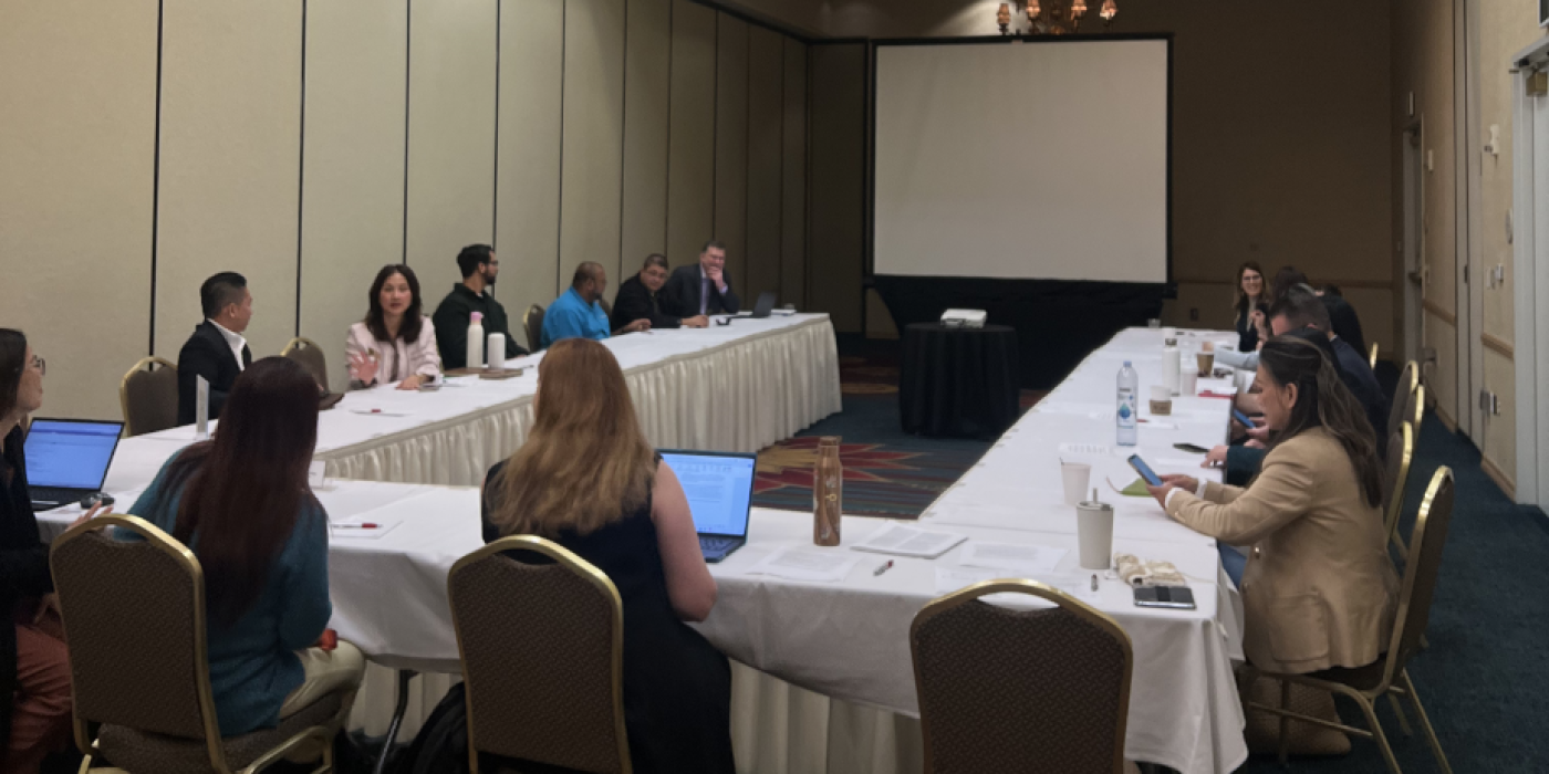 Group of attendees seated at U-shaped conference tables during a formal meeting in a banquet hall, with a projection screen at the front.