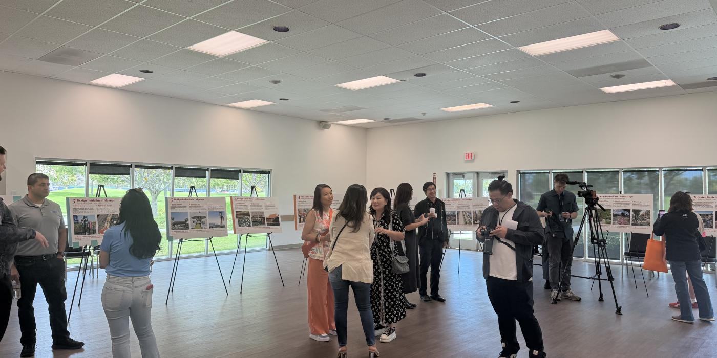 Community members and media gathered at an indoor public meeting, viewing informational display boards on easels.