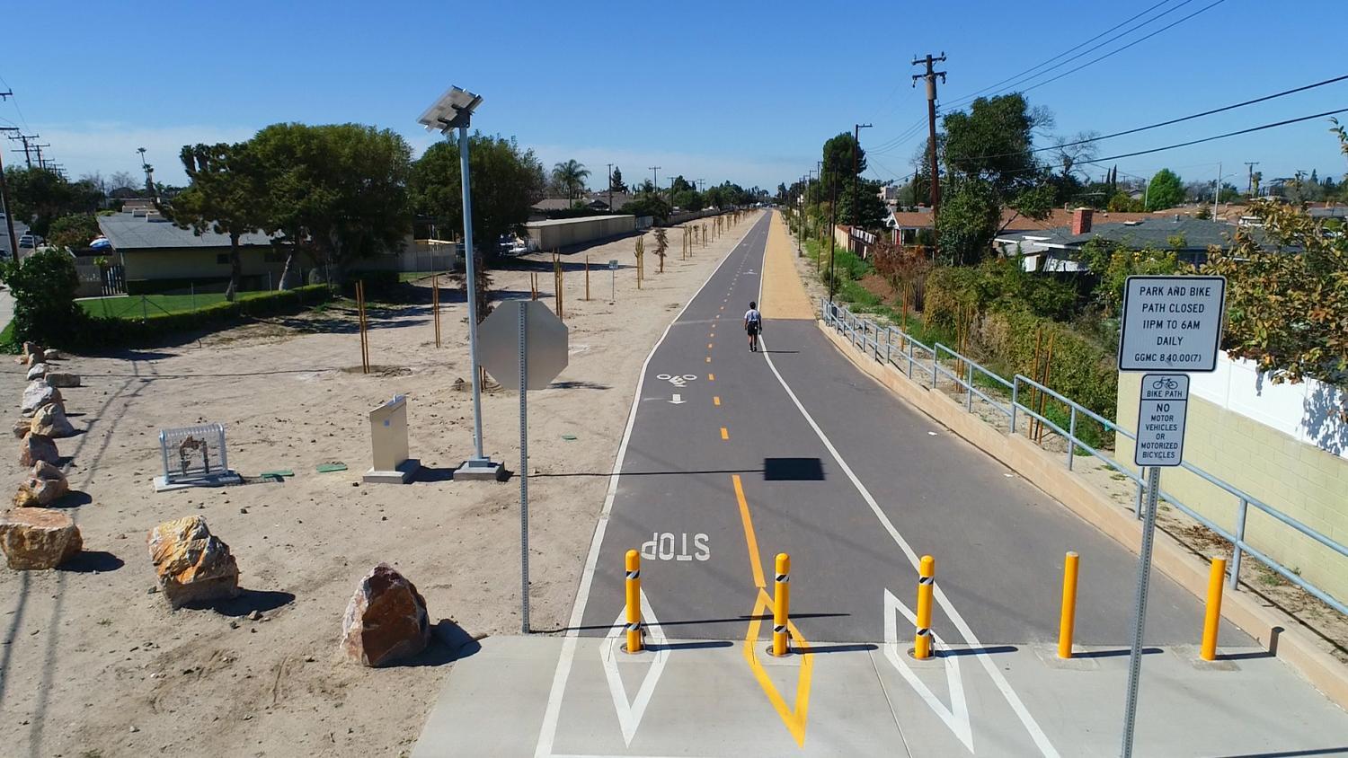 Paved bike path with a cyclist riding along it, STOP marking and bollards at entry, sign reading "Park and Bike Path Closed 11PM to 6AM Daily," solar-powered light pole, and decorative boulders along the path in a residential area of Garden Grove.